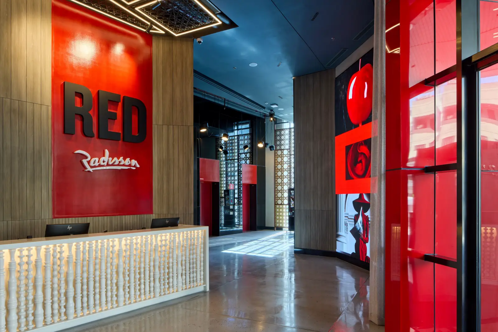 Lobby and reception area of Radisson RED Cebu Mandaue, featuring a bold red wall with the RED Radisson logo, wood panelled accents, a white textured front desk, geometric ceiling lighting and a corridor leading to red toned digital art installations and elevators.