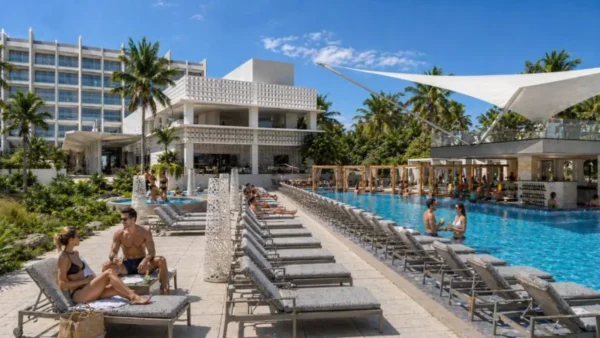 Guests relaxing on sun loungers beside the outdoor pool at Royalton Vessence Barbados, with palm trees, white shade sails, and the resort building in the background