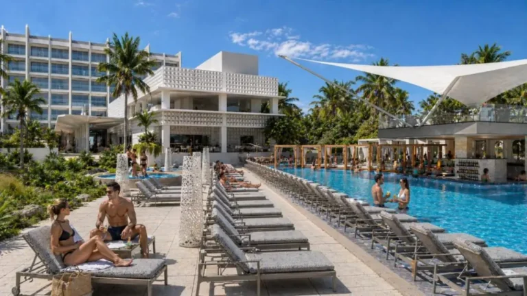 Guests relaxing on sun loungers beside the outdoor pool at Royalton Vessence Barbados, with palm trees, white shade sails, and the resort building in the background