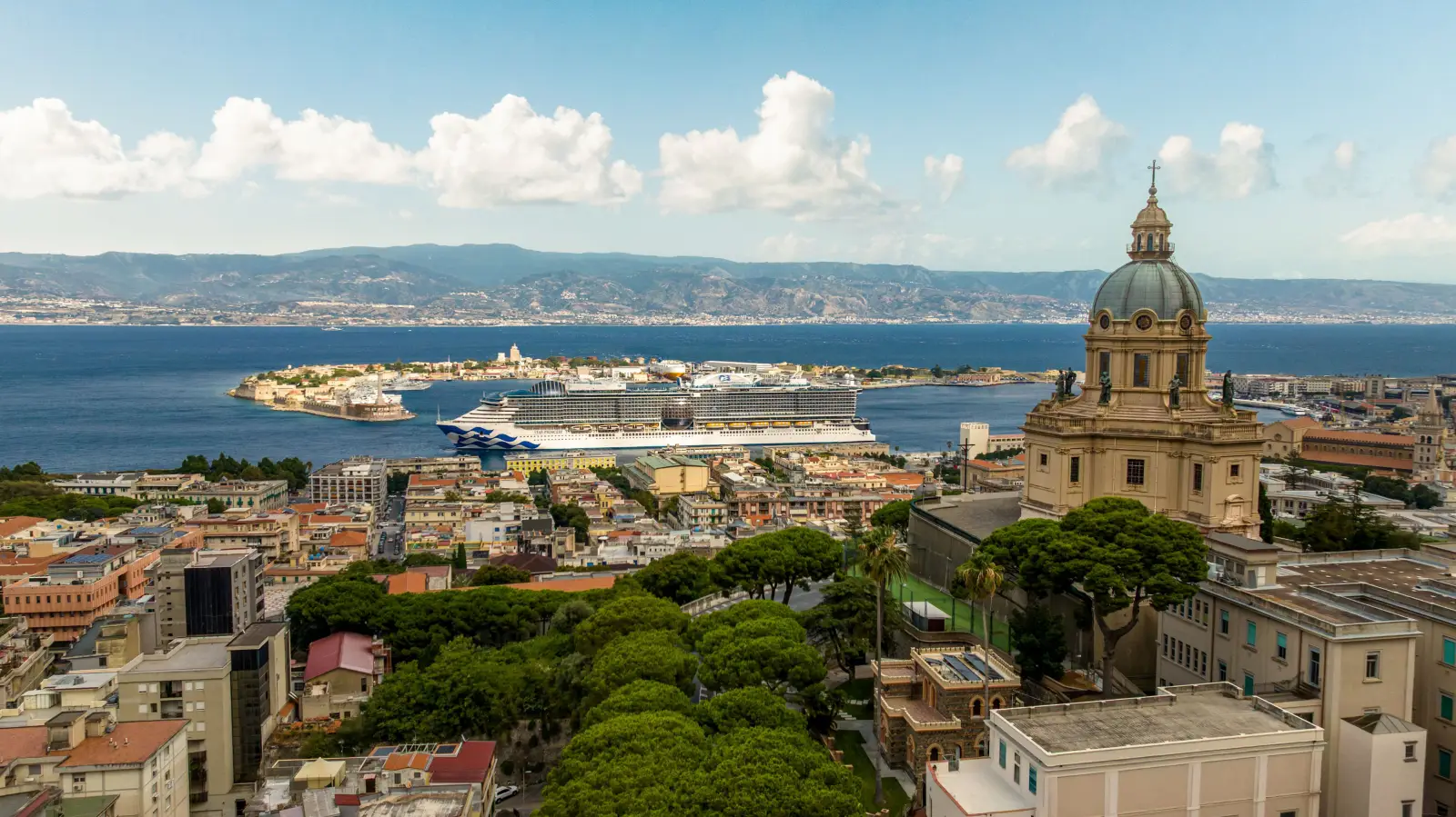 Princess Cruises Is Launching Its Biggest-Ever Europe Season In 2028 3 Aerial view of Messina, Sicily, showing the Sun Princess cruise ship docked at the port beside the Strait of Messina. The foreground features a ornate baroque church dome and lush tree canopy, with the city's low-rise buildings stretching toward the waterfront. The strait and the hills of Calabria on the Italian mainland are visible in the background under a partly cloudy sky.