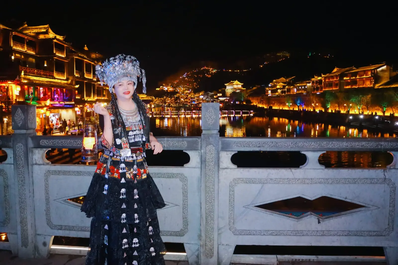Tourist wearing traditional Chinese ethnic costume on a bridge overlooking a brightly lit ancient town at night
