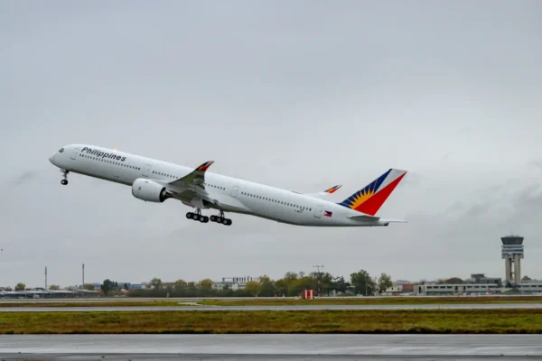 A Philippine Airlines Airbus A350-1000 lifts off from a runway in overcast conditions, showing the airline's distinctive blue, red and yellow sunburst tail livery and "Philippines" fuselage branding, with an air traffic control tower visible in the background