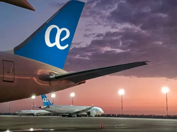 Two Air Europa aircraft parked on the apron at dusk, with a close-up of the blue tail fin bearing the airline's white cursive logo in the foreground and a Boeing 787 Dreamliner visible in the background against a dramatic pink and purple sunset sky