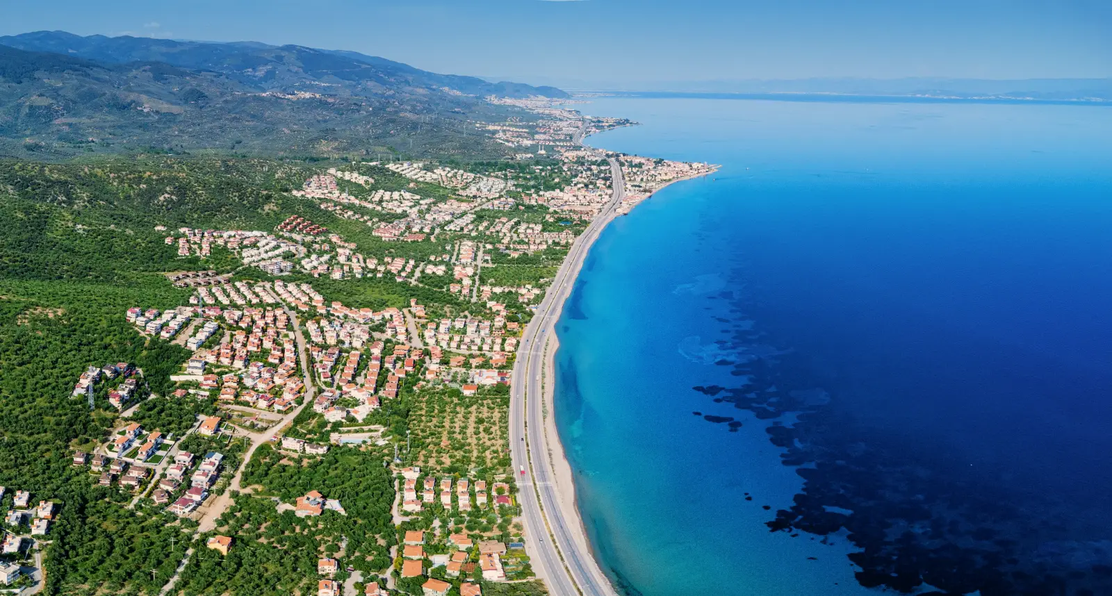 Aerial view of Altinoluk in Edremit, Balikesir, showing the Aegean coastline, residential neighborhoods, green hills and clear blue sea on a sunny day.