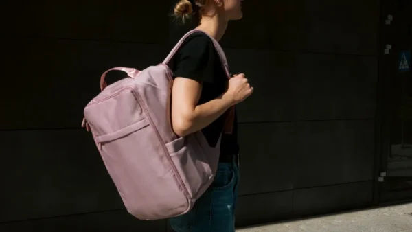 Woman carrying a large pink travel backpack while walking outdoors in casual clothing against a dark urban background.