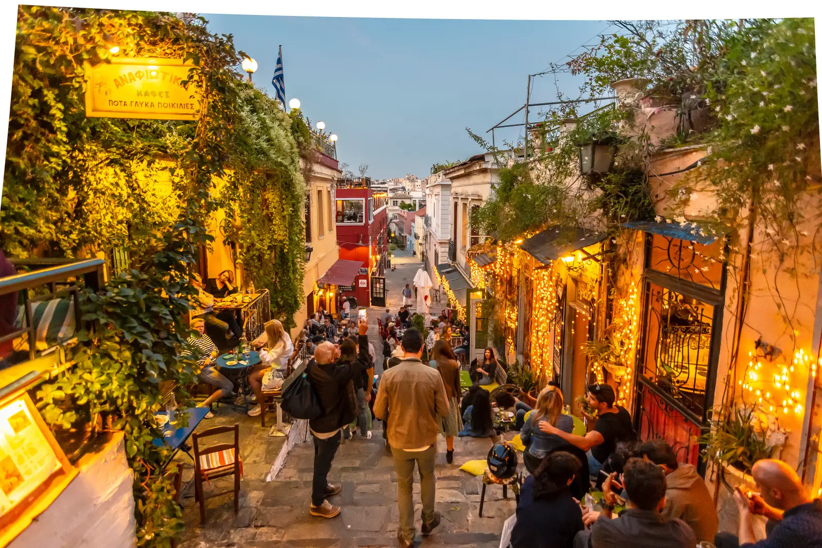 A lively staircase street in the Plaka district of Athens at dusk, lined with warmly lit cafes, string lights, and climbing vines, with groups of visitors and locals seated at outdoor tables on both sides