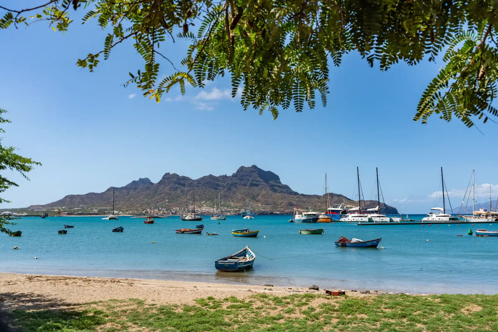 Fishing boats and sailing yachts anchored in a calm turquoise bay with a rugged volcanic mountain in the background, Cape Verde