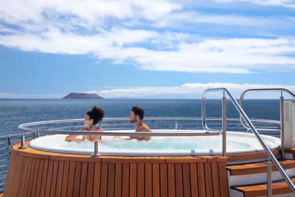 Two guests relaxing in the outdoor deck jacuzzi aboard Celebrity Flora, surrounded by teak wood panelling, with the calm blue waters of the Galapagos archipelago and a distant volcanic island visible under a bright sky
