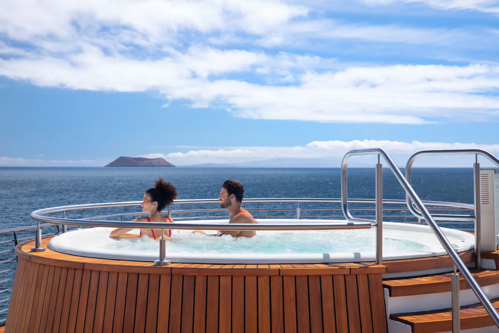Celebrity Cruises’ 2028 Galapagos Expeditions Offer Once-In-A-Lifetime Adventures 2 Two guests relaxing in the outdoor deck jacuzzi aboard Celebrity Flora, surrounded by teak wood panelling, with the calm blue waters of the Galapagos archipelago and a distant volcanic island visible under a bright sky