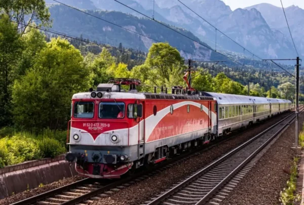 A red and white CFR Calatori electric locomotive pulls a passenger train along a curved track through a forested mountain valley, with electrified overhead lines and steep rocky peaks in the background.