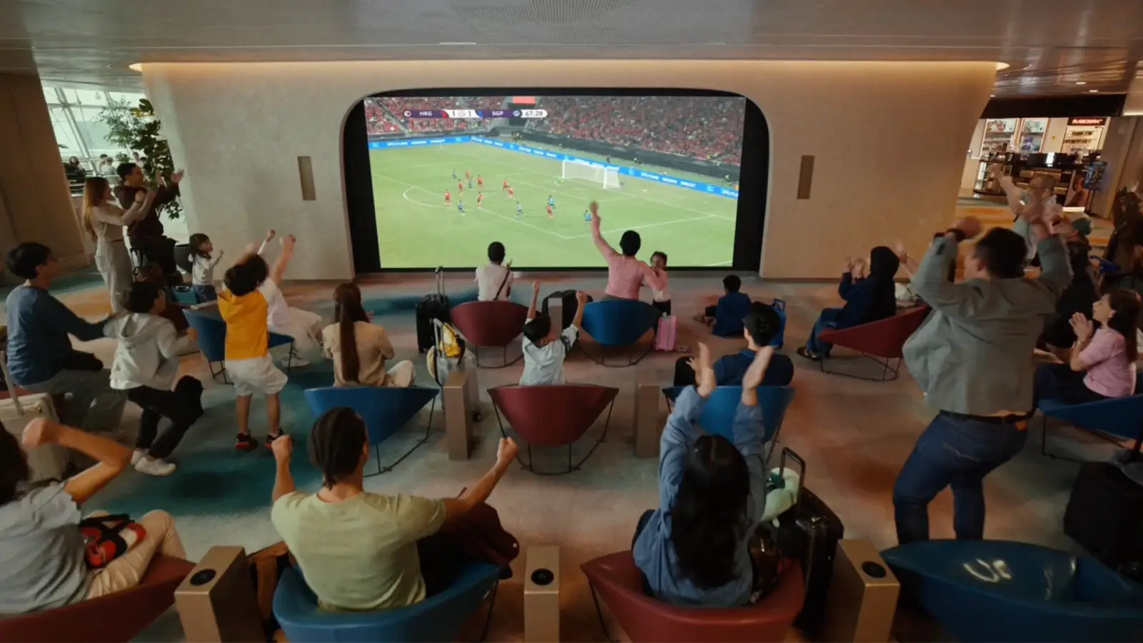Changi Airport Kicks Off Football Fever With New Film, Free Match Screenings And Airport-Wide Activities 4 Excited football fans raising their arms and cheering while watching a live football match between Hong Kong and Singapore on a large curved screen at a Changi Airport TV lounge, seated on blue and red chairs with luggage nearby and retail shops visible in the background