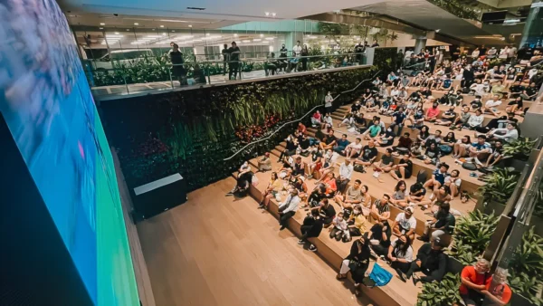 Home 2 Large crowd seated on tiered wooden steps watching a live football match on a big screen at ST3PS in Changi Airport Terminal 3, with a green living plant wall behind them, spectators also watching from a glass-railed upper level mezzanine, and tropical plants framing the space
