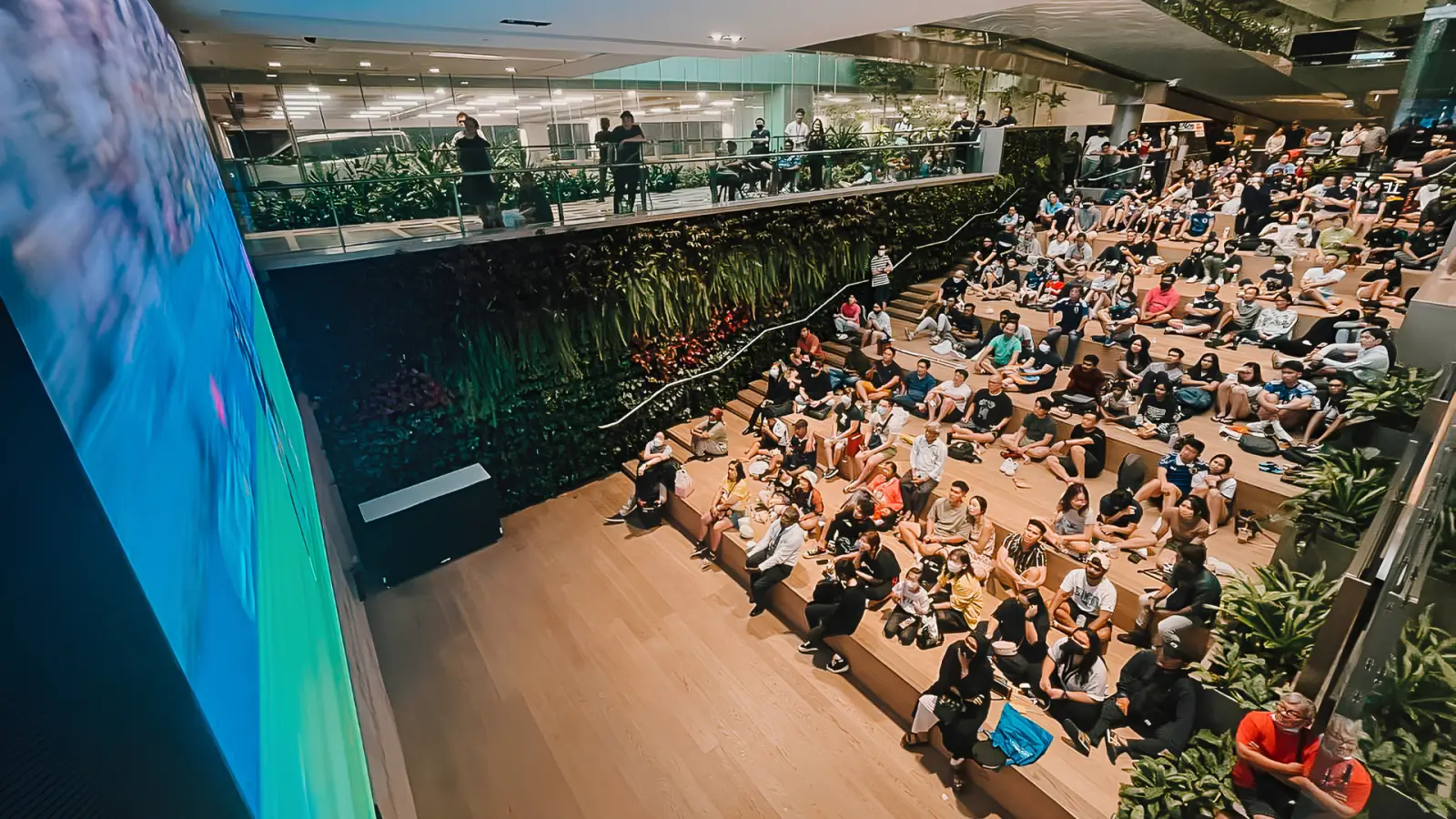 Changi Airport Kicks Off Football Fever With New Film, Free Match Screenings And Airport-Wide Activities 4 Large crowd seated on tiered wooden steps watching a live football match on a big screen at ST3PS in Changi Airport Terminal 3, with a green living plant wall behind them, spectators also watching from a glass-railed upper level mezzanine, and tropical plants framing the space