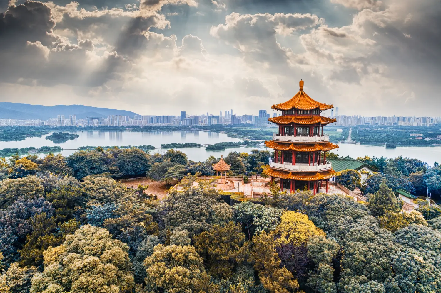 Aerial view of a multi-tiered traditional Chinese pagoda with golden rooftops and red pillars, surrounded by dense green trees, with a wide lake and modern city skyline visible in the background under a dramatic cloudy sky.