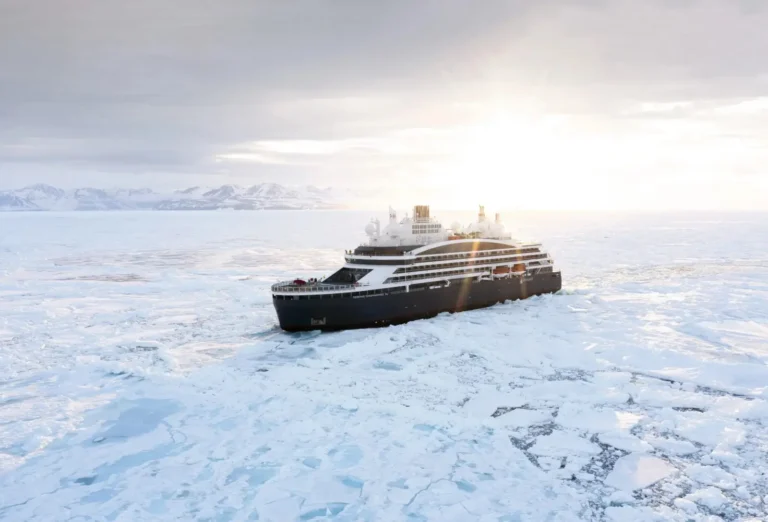 Cruise Travel 22 The luxury icebreaker Le Commandant Charcot cutting through Arctic pack ice off Greenland, with snow-covered mountains and a low sun on the horizon