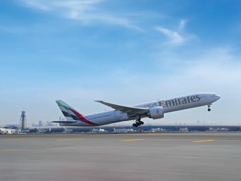An Emirates Boeing 777 lifting off from Dubai International Airport runway with the Dubai skyline and airport control tower visible in the background under a clear blue sky
