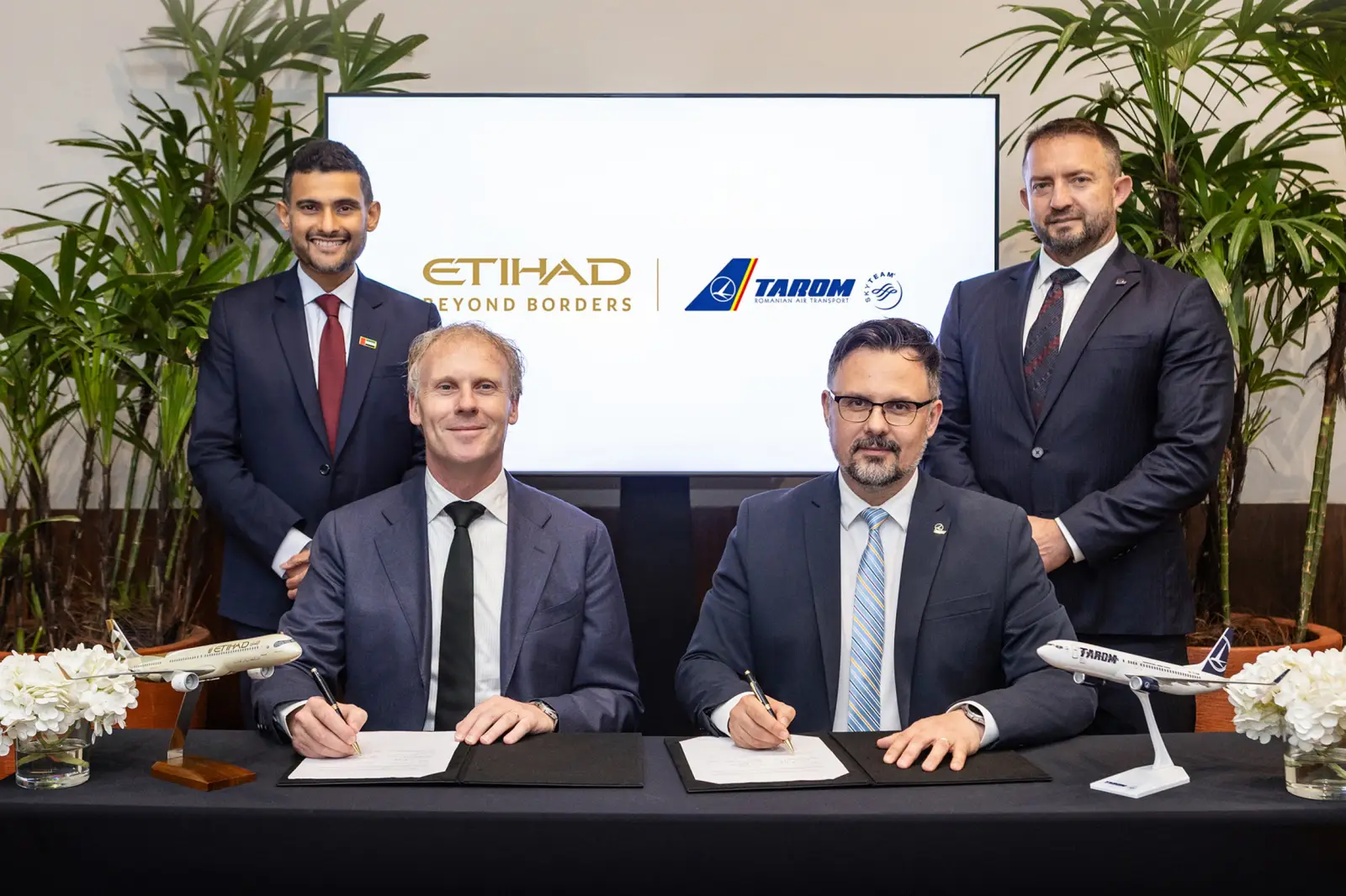 Etihad Codeshare With Tarom Connects Abu Dhabi To 11 Romania And Eastern Europe Destinations 3 Four airline executives at a signing ceremony for the Etihad Airways and TAROM codeshare agreement, two seated at a table signing documents with aircraft models in front of them and the Etihad and TAROM logos displayed on a screen behind them.