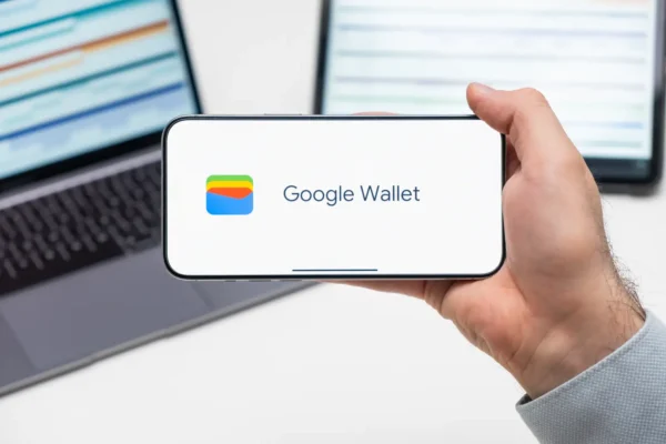 A person in a grey suit jacket holds a smartphone horizontally displaying the Google Wallet app logo and name on a white screen, with a blurred laptop and tablet visible in the background on a white desk