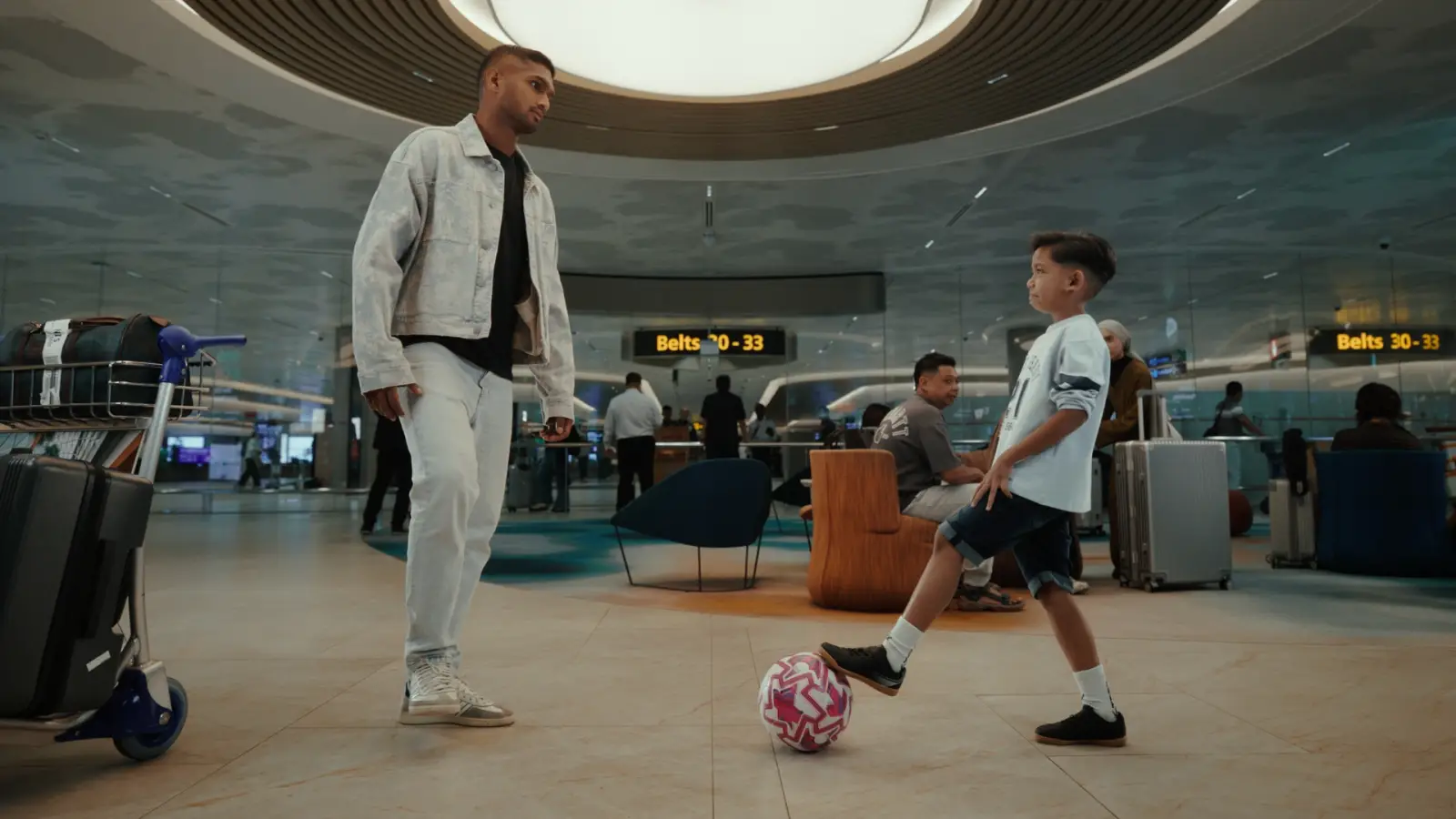 Changi Airport Kicks Off Football Fever With New Film, Free Match Screenings And Airport-Wide Activities 3 Singapore national football team captain Hariss Harun in a denim jacket passing a pink and white football to a young boy in the arrivals baggage hall of Changi Airport, with yellow belt number displays, luggage trolleys, and travellers visible in the background under a circular ceiling light