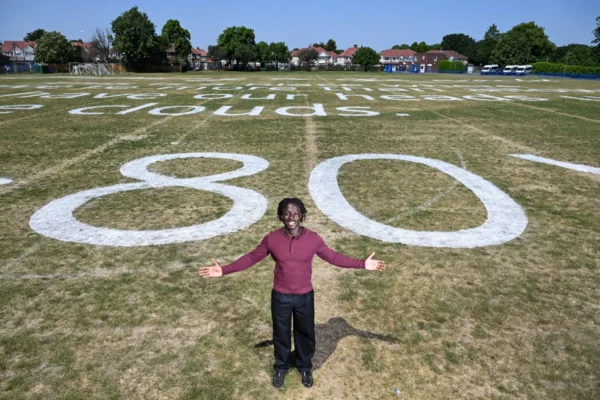 Poet David Larbi stands in front of a giant "80" painted on a grass field, with poem verses written across the ground behind him.