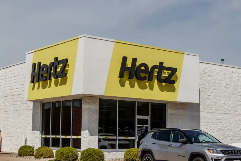 Exterior view of a Hertz car rental branch with yellow branding and an SUV parked outside.
