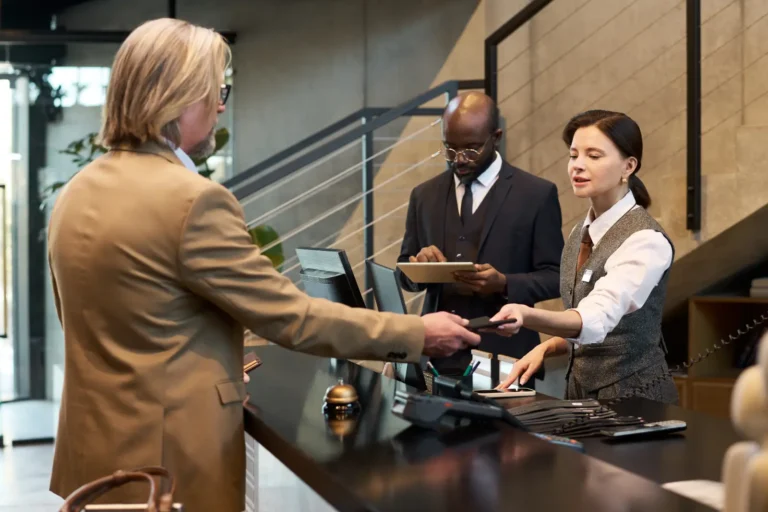 Hotel front desk staff assisting a guest at check-in while a manager reviews a tablet, with multiple devices visible on the reception counter