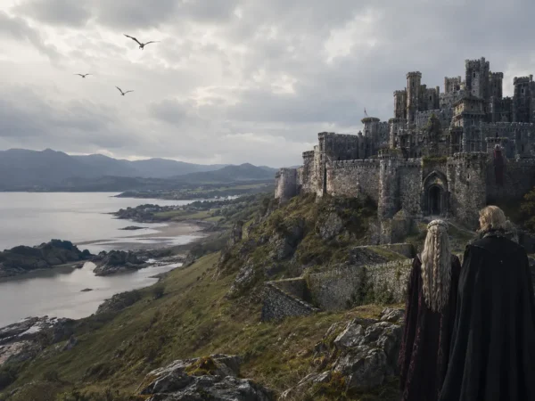 Fantasy-inspired medieval castle overlooking a dramatic Welsh-style coastline with mountains, cliffs and two cloaked figures in the foreground
