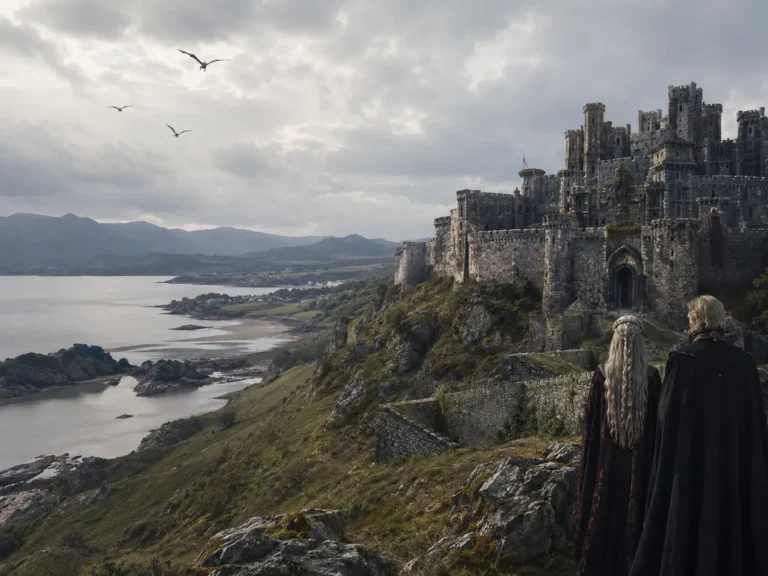 Tours 20 Fantasy-inspired medieval castle overlooking a dramatic Welsh-style coastline with mountains, cliffs and two cloaked figures in the foreground