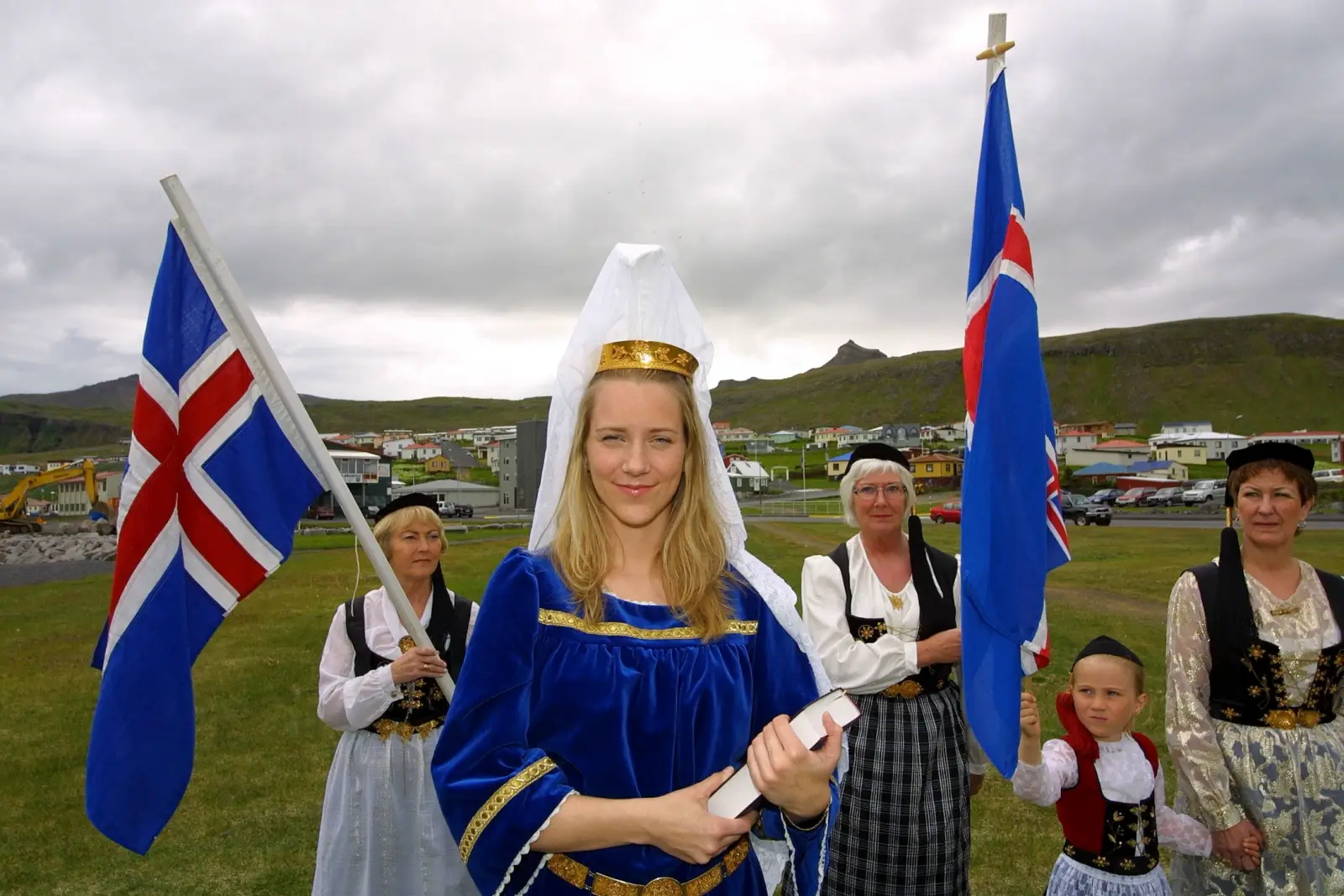 A young woman dressed as Iceland's Fjallkonan, wearing a blue velvet gown, gold crown and white veil, stands holding a scroll during a National Day celebration in Olafsvik, with Icelandic flags and women in traditional folk costumes behind her, against a backdrop of coastal hills and the town.