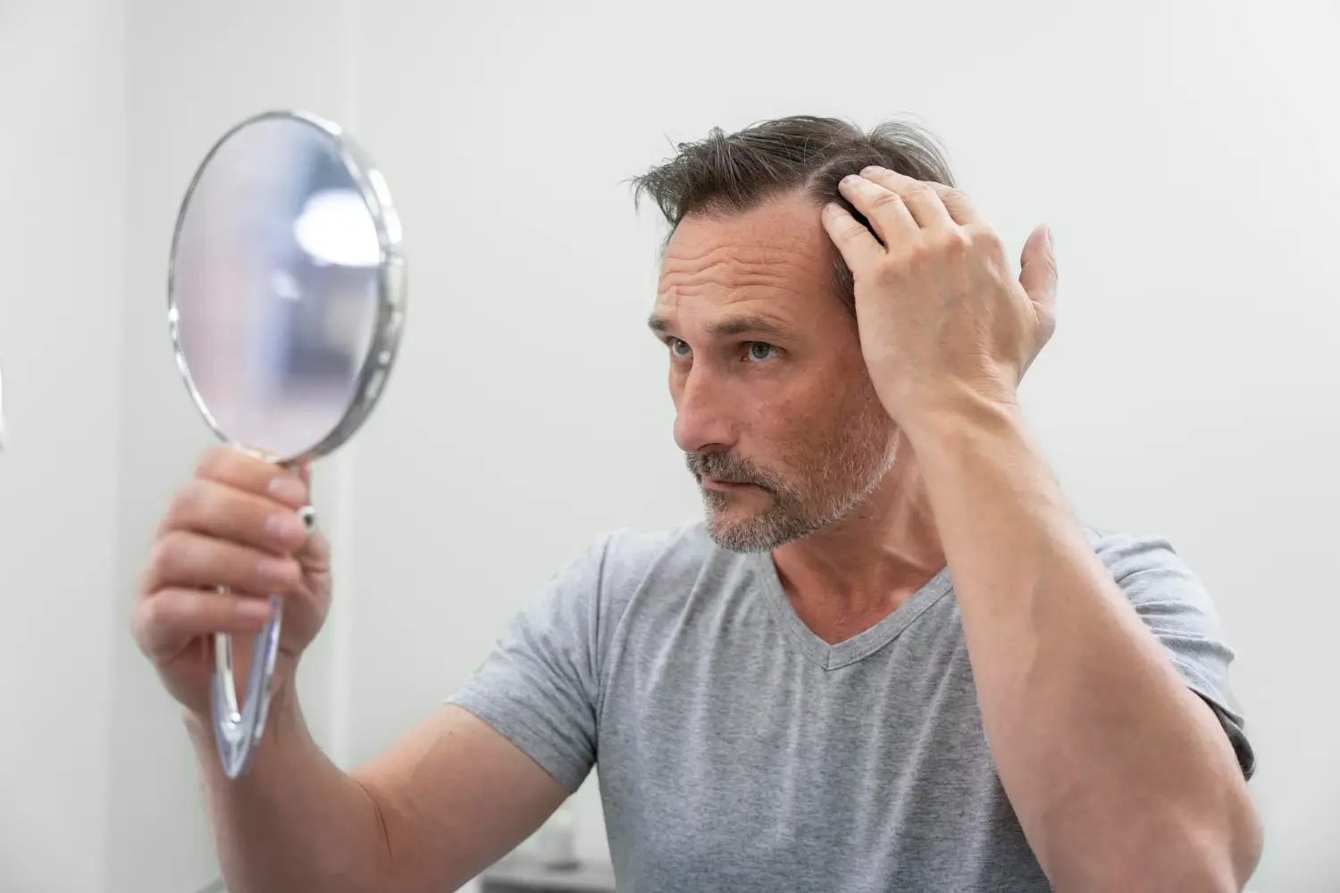Middle-aged man checking his hairline in a mirror while examining signs of hair loss at home.