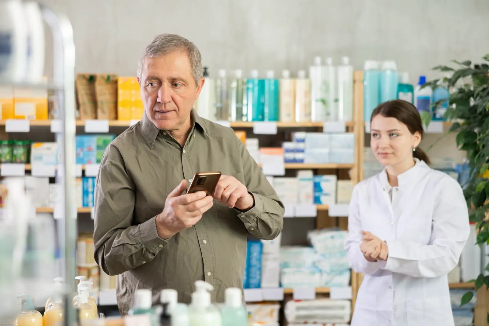 An older man in a pharmacy holds up his smartphone, comparing information while a female pharmacist in a white coat stands nearby, with shelves of medicines and toiletries in the background.