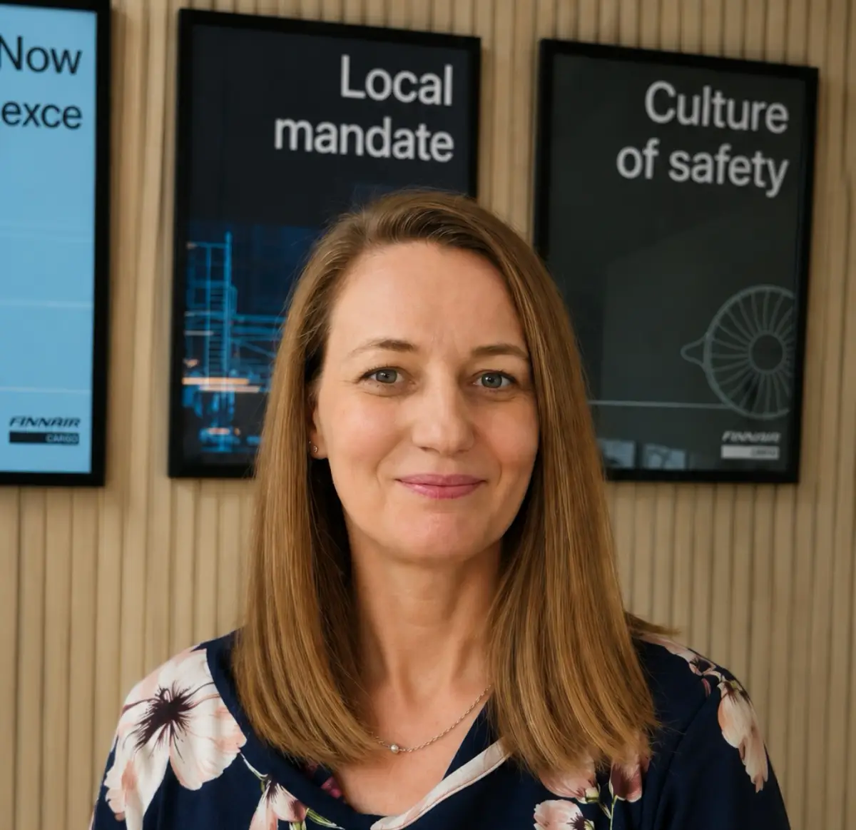 Headshot of Minna Virmajoki, smiling and wearing a floral top, standing in front of wood paneled wall with Finnair branded posters reading Local Mandate and Culture of Safety.