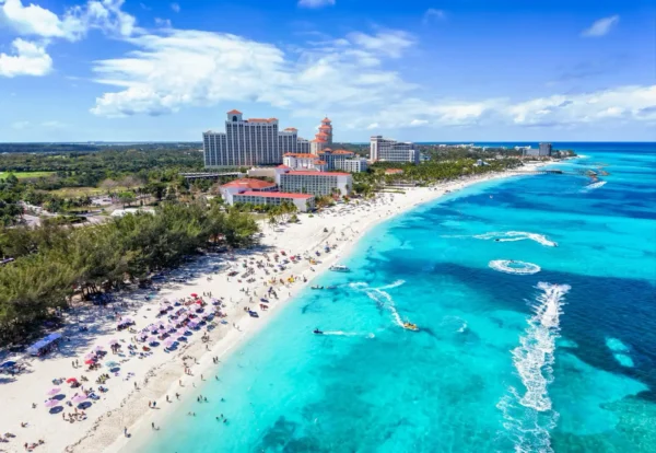 Home 2 Aerial view of Goodman's Bay public beach in Nassau, Bahamas, showing a crowded white sand beach with umbrellas and sunbathers, resort towers along the coastline, and several jet skis leaving wake trails across the turquoise water