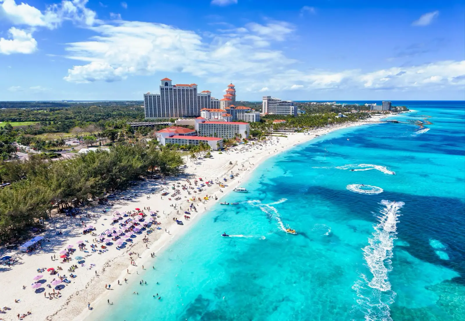 Why The Us Embassy Just Issued A Bahamas Beach Warning 4 Aerial view of Goodman's Bay public beach in Nassau, Bahamas, showing a crowded white sand beach with umbrellas and sunbathers, resort towers along the coastline, and several jet skis leaving wake trails across the turquoise water