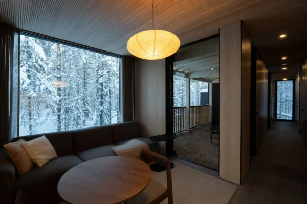 Home 4 Warm interior of a NOA Villas private villa in Finnish Lapland featuring a dark sofa with cream cushions, a round wooden dining table, a glowing round paper pendant lamp, and large floor-to-ceiling windows overlooking a snow-covered Arctic birch forest, with an open glass door leading to a wooden balcony