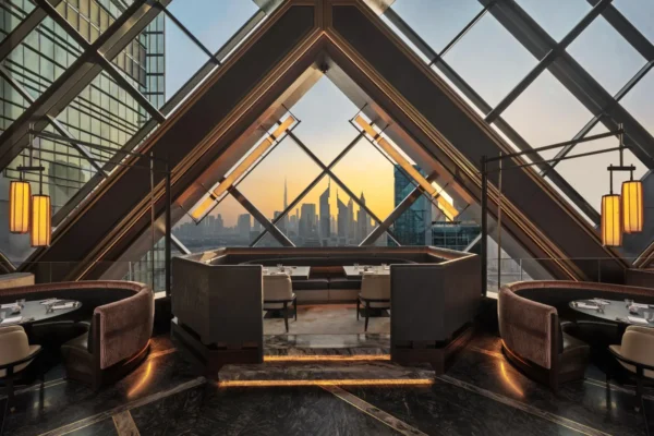 Interior of Nobu One Za'abeel restaurant in Dubai, showing the dramatic crisscrossing geometric glass facade framing a panoramic golden-hour view of the Dubai skyline including the Burj Khalifa, with dark marble floors, warm pendant lighting, and upholstered booth seating in the foreground
