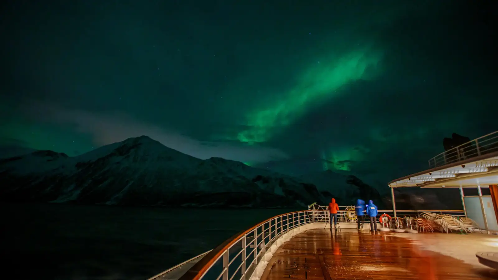Northern Lights In Sight: Expedition Cruises To Scandinavia In 2026/27 With Hapag-Lloyd Cruises 3 Guests watching the green aurora borealis from the open deck of HANSEATIC nature off a snow-covered Norwegian fjord