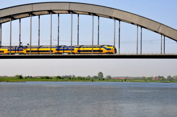A yellow and blue Nederlandse Spoorwegen double-decker train crossing an arch bridge over a Dutch river with flat green countryside in the background
