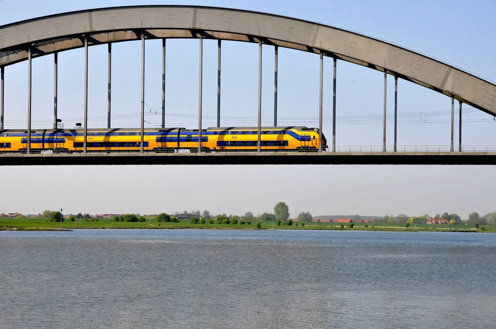 A yellow and blue Nederlandse Spoorwegen double-decker train crossing an arch bridge over a Dutch river with flat green countryside in the background