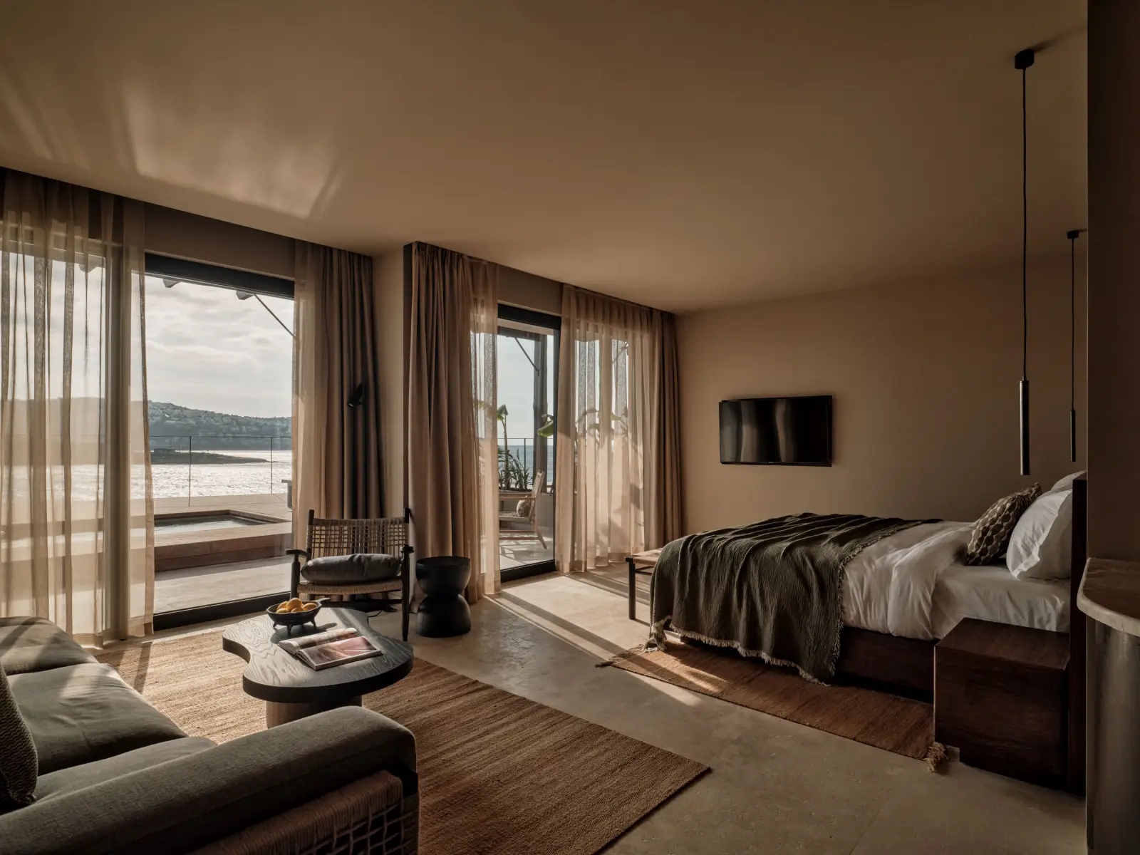 A spacious hotel room at OKU Bodrum featuring a king bed with olive green and white linens, a seating area with sofas and a round wooden coffee table, and floor to ceiling windows opening onto a private terrace with sea views.