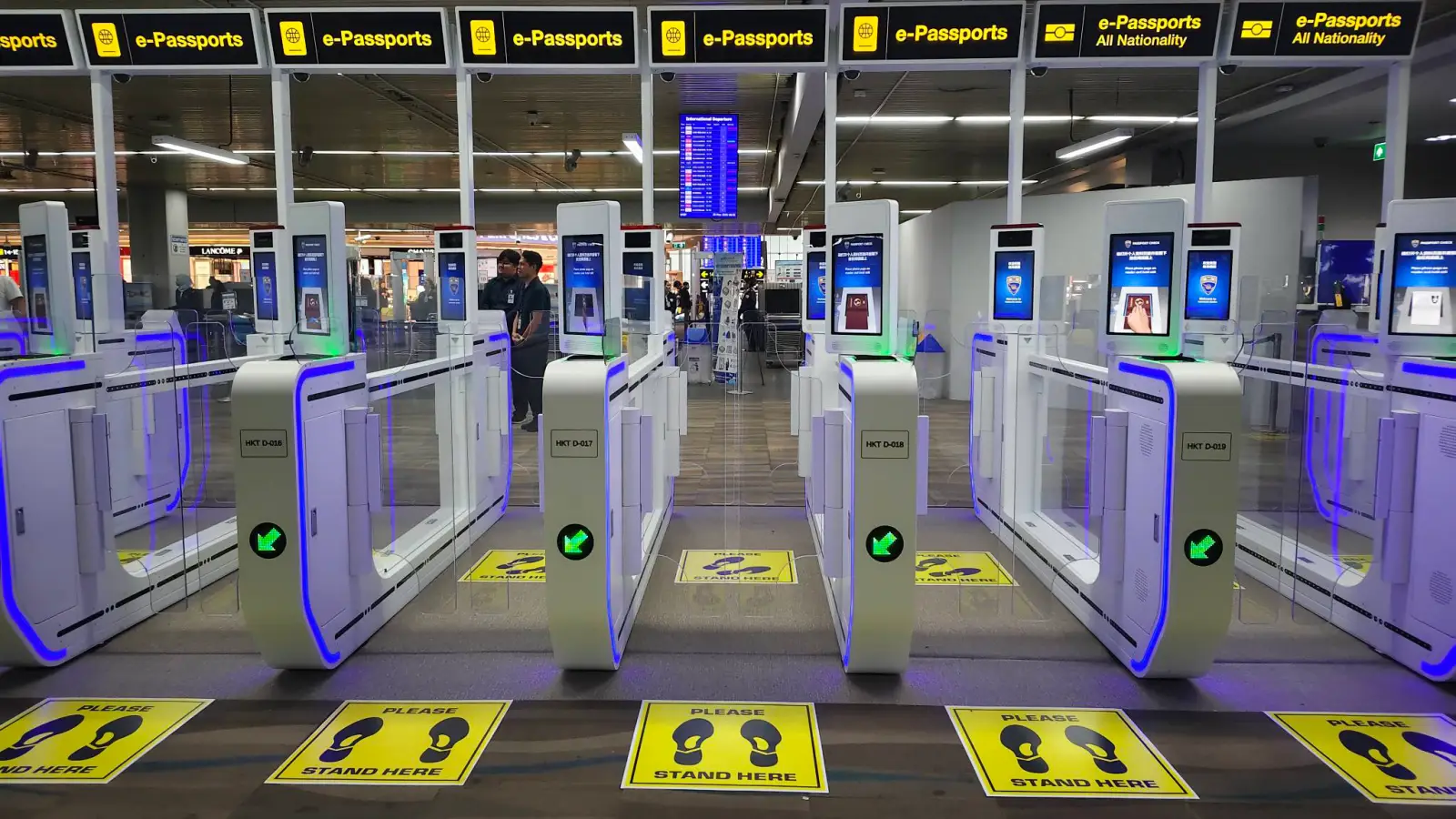 A row of automated border control gates with blue LED lighting at Phuket International Airport, with yellow overhead signs reading e-Passports and e-Passports All Nationality, yellow floor markers indicating where passengers should stand, and an international departures flight information board visible in the background.