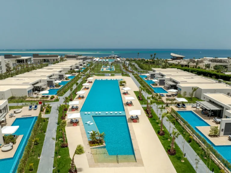 Aerial view of Rixos Premium Magawish Bay View in Hurghada, showing multiple swimming pools flanked by private villas, sun loungers, landscaped gardens, and the Red Sea in the background.