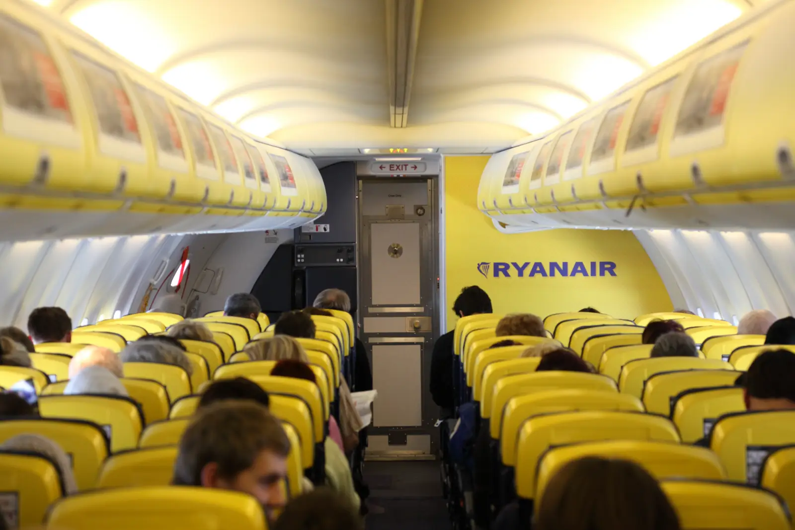 Interior of a Ryanair aircraft cabin with passengers seated and yellow seats facing the rear exit