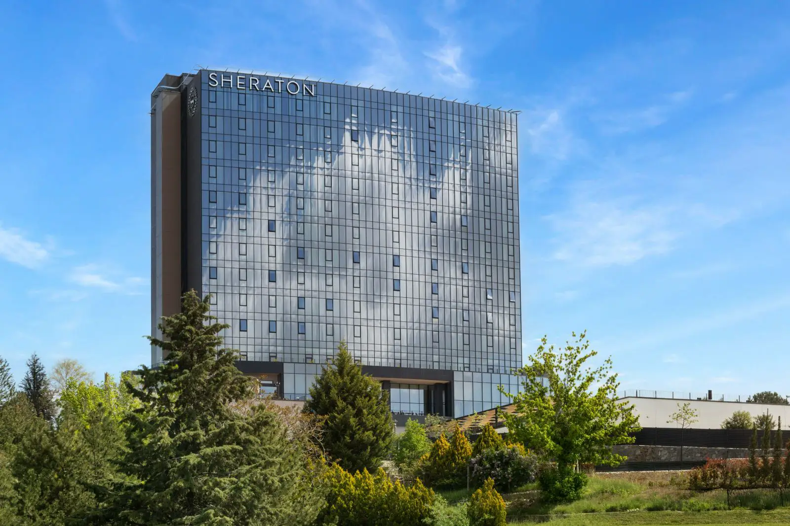 Sheraton Uşak Hotel And Thermal Spa Opens As Marriott'S 500Th Emea Premium Hotel 3 A tall glass fronted building displaying the Sheraton sign at the top, set against a clear blue sky with trees and landscaped grounds in the foreground, showing the exterior of the Sheraton Usak Hotel and Thermal Spa in Turkiye.
