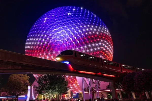 The Spaceship Earth geodesic sphere at EPCOT, Walt Disney World, illuminated at night in patriotic red, white and blue lighting. A Disney monorail glides along the elevated track in the foreground, with trees and a crowd of guests gathered below under the glowing landmark.
