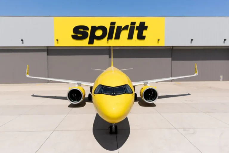 A yellow Spirit Airlines narrow-body aircraft parked on the tarmac directly in front of a large hangar bearing the Spirit logo in black lettering on a yellow background, viewed head-on under a clear blue sky.