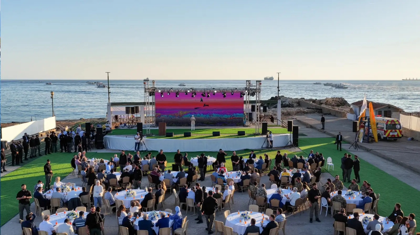Syria Opens Its 2026 Summer Tourism Season On The Mediterranean Coast 1 Aerial view of the official 2026 Syria summer tourism season launch event held on the Mediterranean waterfront in Tartus, with guests seated at round tables facing a large LED stage, and fishing boats visible on the sea in the background