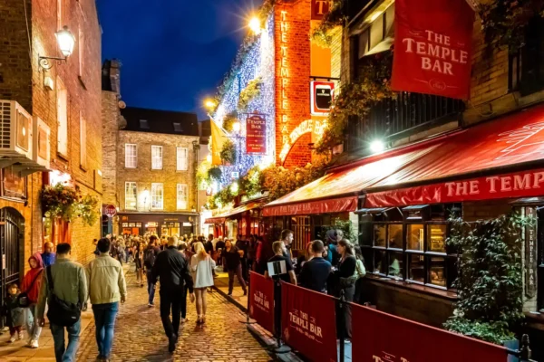 The Temple Bar pub lit up at night in Dublin, with crowds walking the cobblestone street past its red facade and string lights