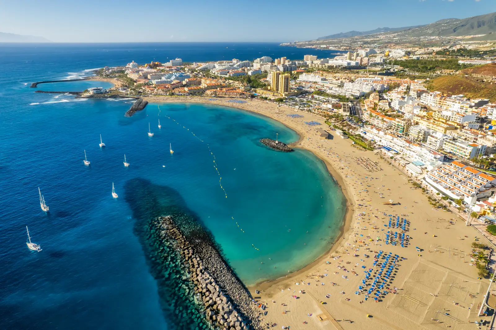 Aerial view of Playa de Las Vistas in Los Cristianos, Tenerife, showing a curved sandy beach with blue sunbeds, turquoise sheltered water, sailing boats anchored offshore, a rock breakwater, and a dense resort townscape stretching along the coastline with mountains in the background