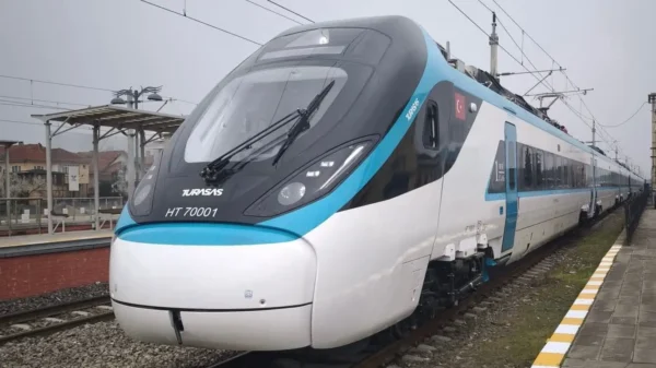 A white and turquoise TURASAS-branded high-speed train unit numbered HT 70001 stands at a platform on a grey overcast day, with overhead electrification cables visible and residential buildings in the background